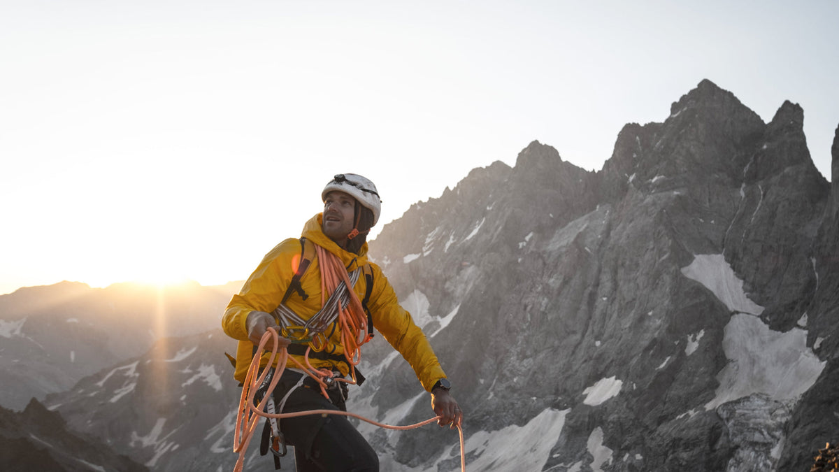 Alpiniste en veste jaune, corde lovée à l'épaule, face aux aiguilles enneigées au lever du soleil