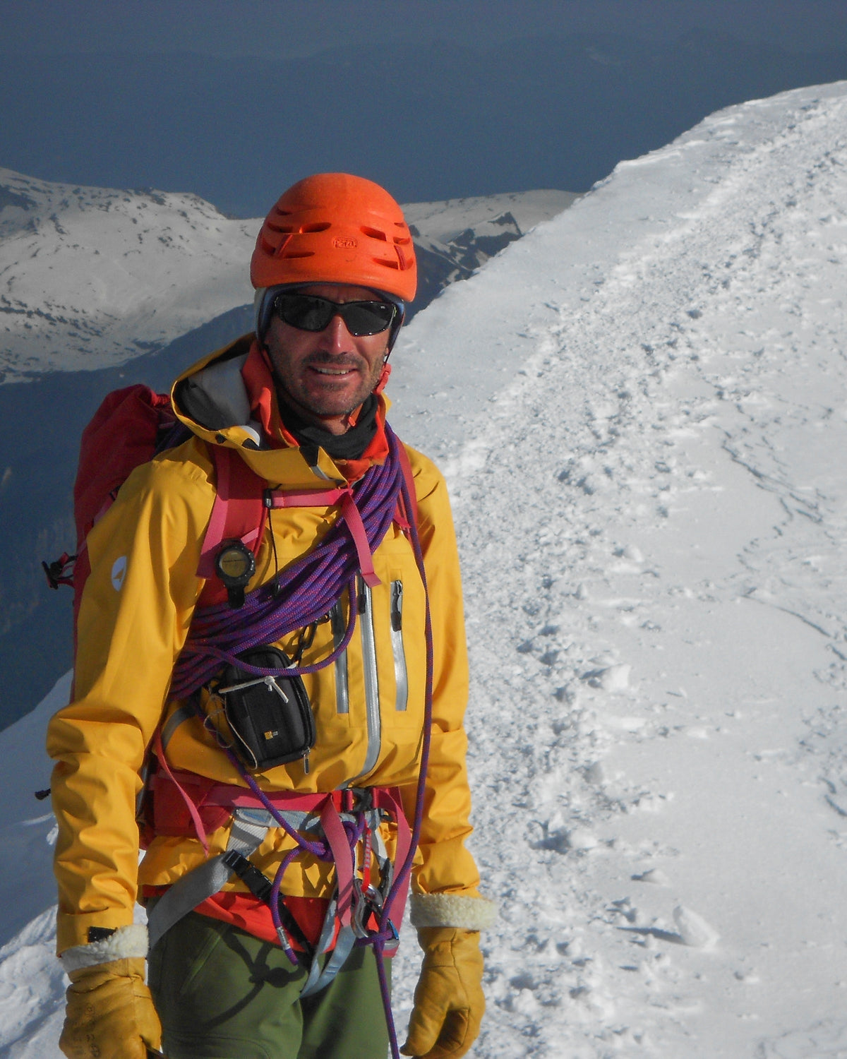 Alpiniste casqué souriant sur une arête enneigée, corde et matériel au baudrier