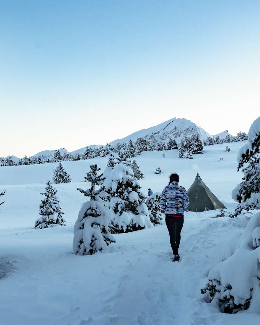 Randonneur rejoignant une tente de bivouac entre des sapins enneigés