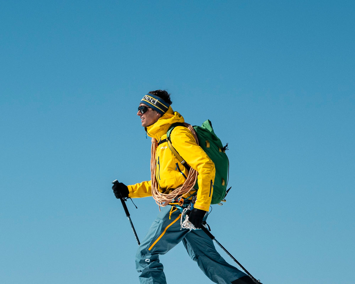 Homme en ski de randonnée, grande foulée en peaux corde à l'épaule, portant le bandeau Lagoped GHEADBAND 70 anthracite sur fond de ciel