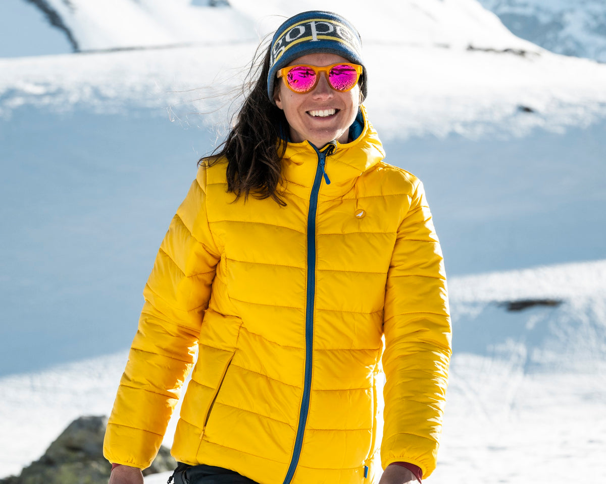 Femme en randonnée dans la neige portant la doudoune à capuche Lagoped HEYDO2 Hoodie jaune soleil, marche face aux sommets enneigés