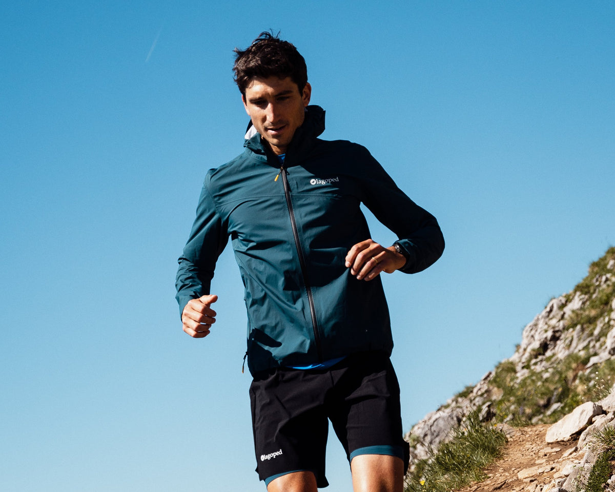 Homme en trail running sur un sentier de montagne portant la veste coupe-vent Lagoped NAGALAKA bleu sarcelle, en été